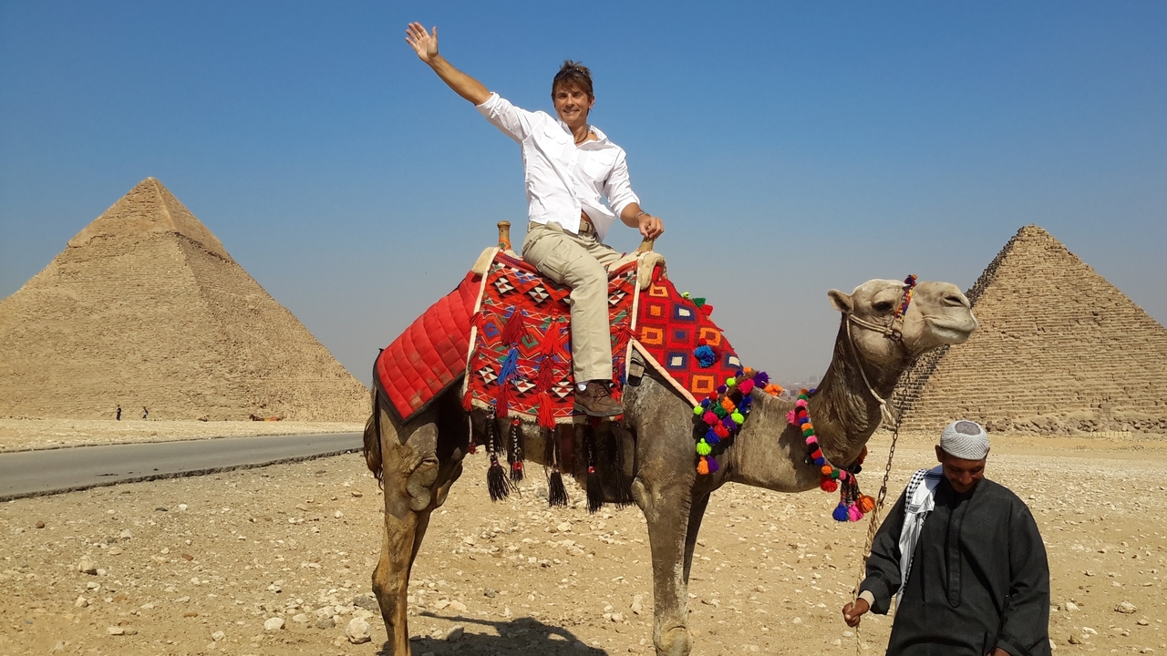 Un homme à dos de chameau avec des pyramides en arrière-plan.