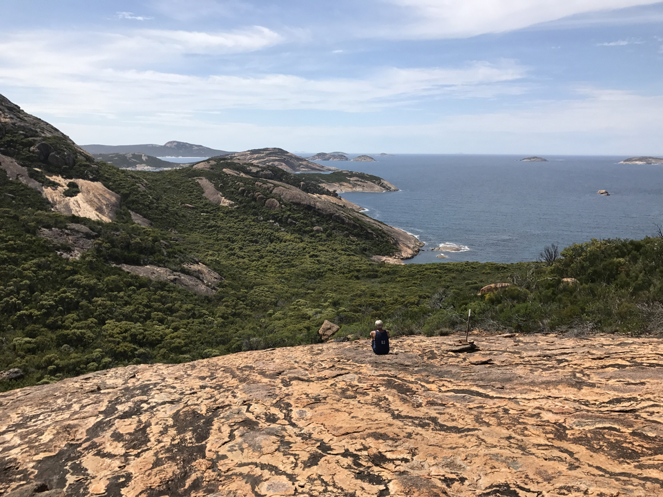A lone hiker sits on a rocky outcrop overlooking green hills that tumble into a rugged turquoise coastline and distant islands.