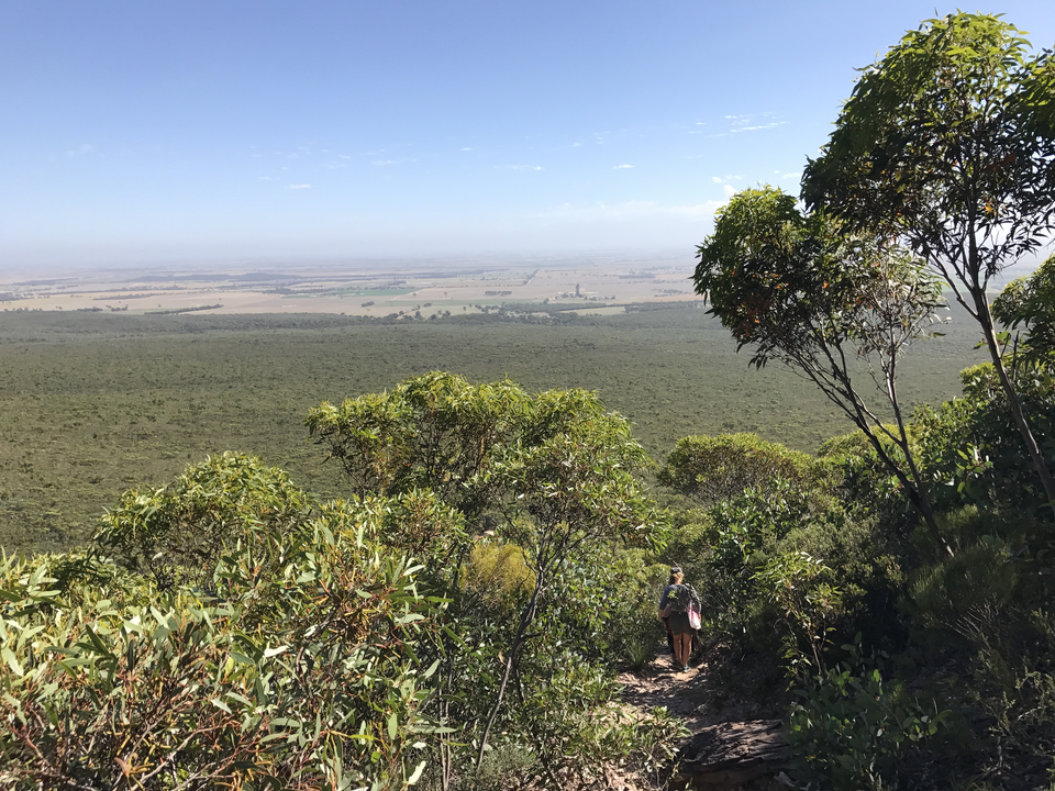 Hiker stands on a mountain trail overlooking vast plains and farmland stretching to the horizon.