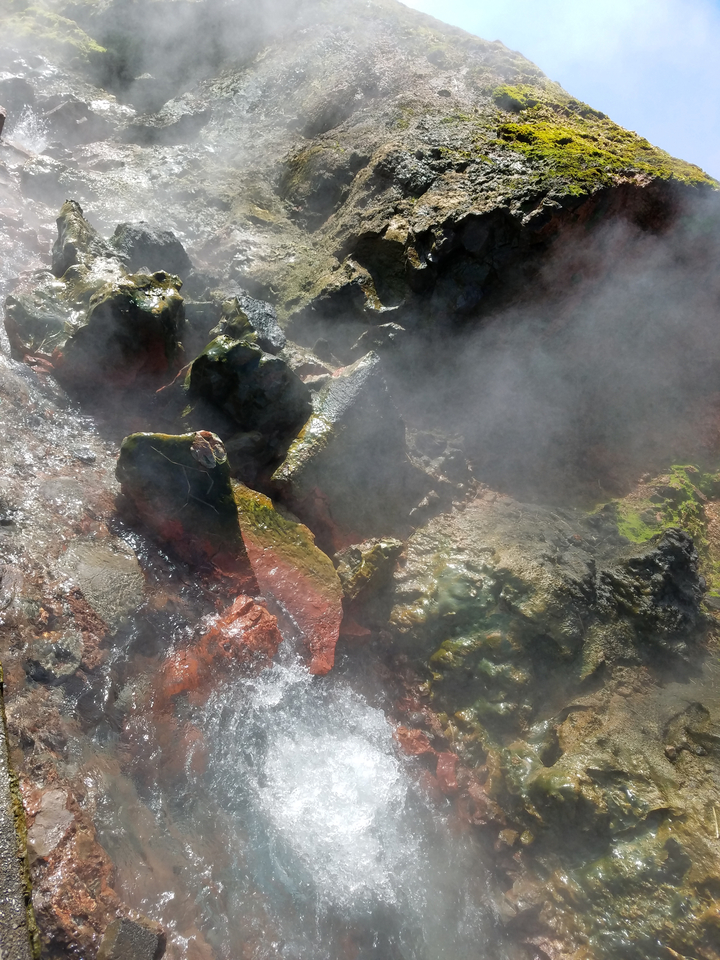 Steam rises over colourful mineral-stained rocks at a geothermal area.