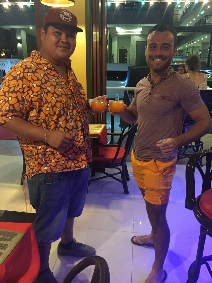 Two men in matching orange tones clinking tropical cocktails inside a bar