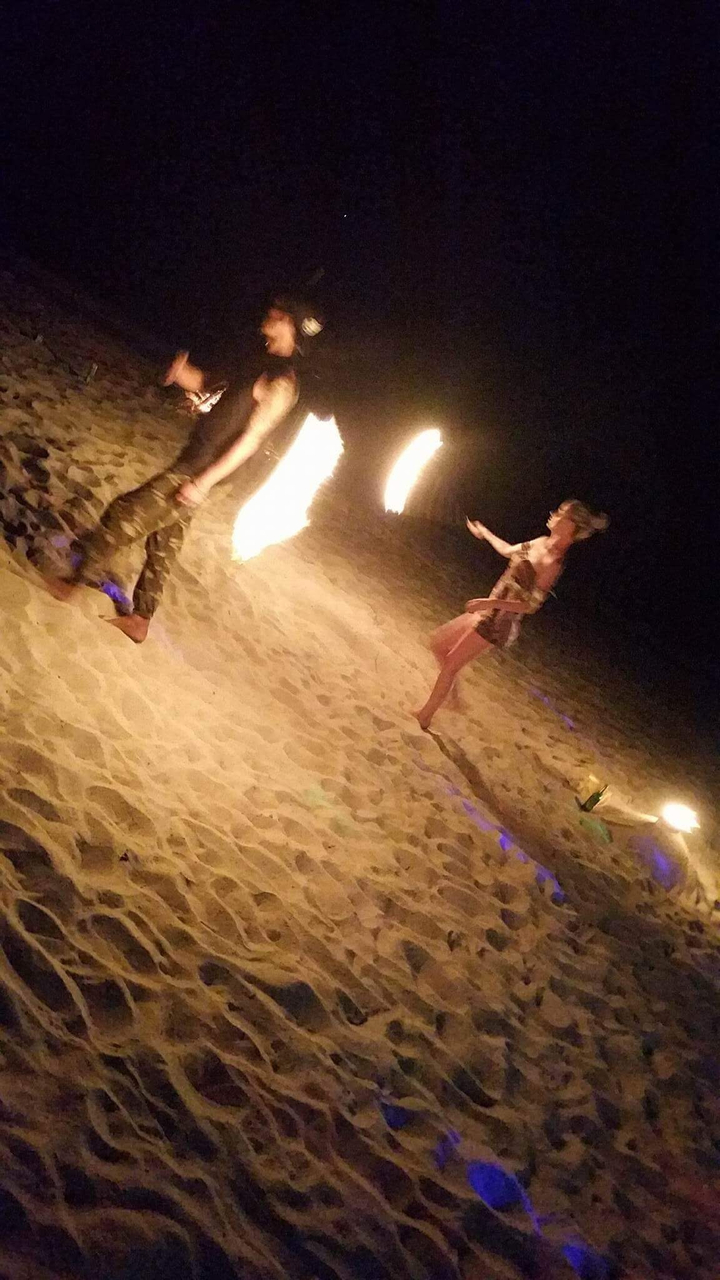 Nighttime beach fire performance with a woman catching flaming baton on sandy shore.