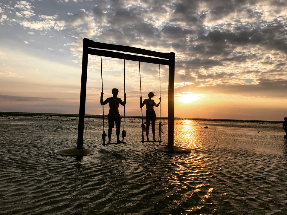 Silhouettes of people on swings at sunset over water.