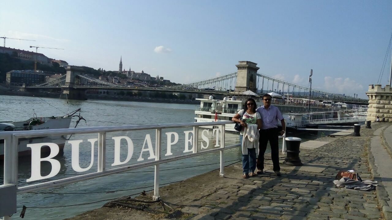 Deux personnes posant près du panneau Budapest, avec une rivière et un pont en arrière-plan.