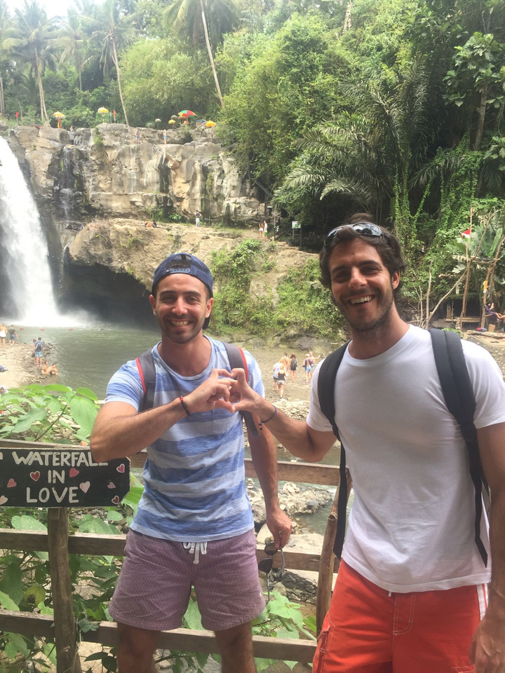 Two smiling travellers making a heart gesture with hands in front of a jungle waterfall.