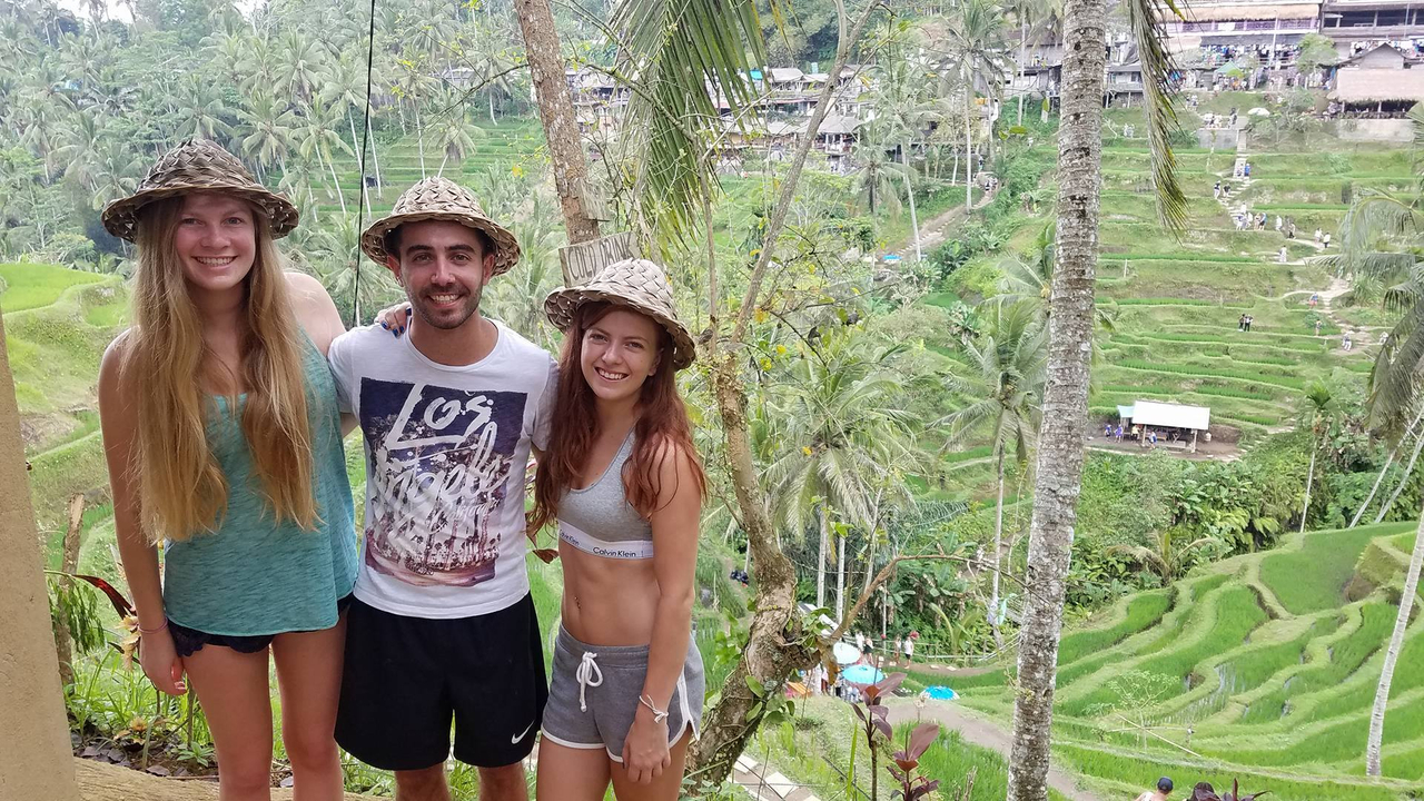 Three friends wearing straw hats smiling on a balcony with sweeping views of emerald rice terraces.