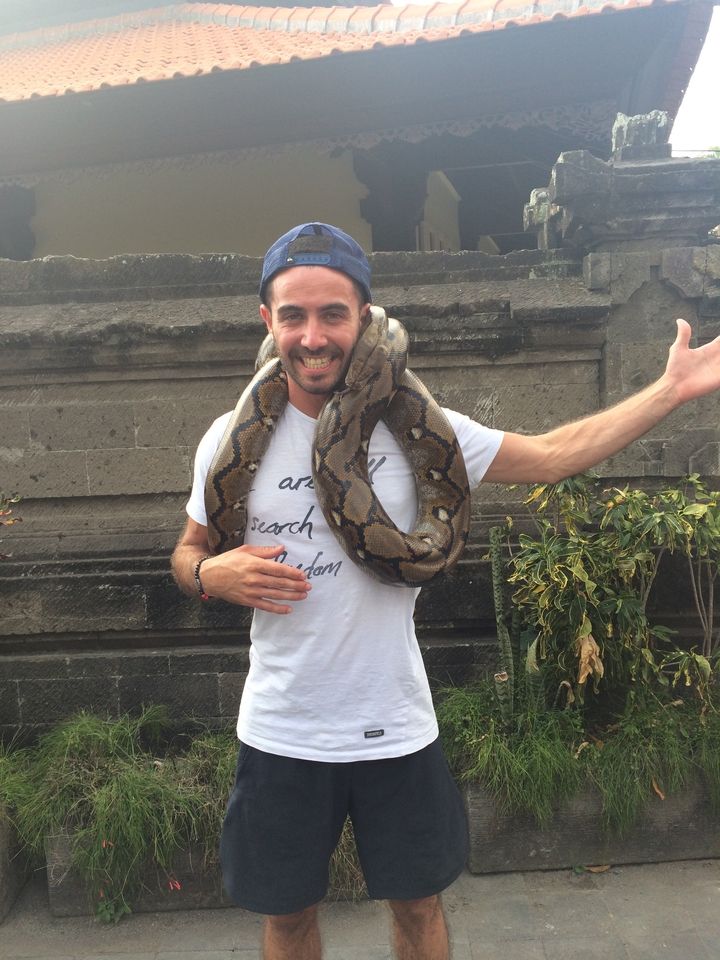 Smiling traveller with a python draped around neck posing against a stone wall.