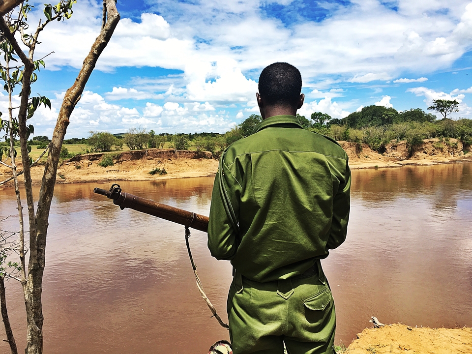 Personne debout au bord d'une rivière, un fusil sur l'épaule.