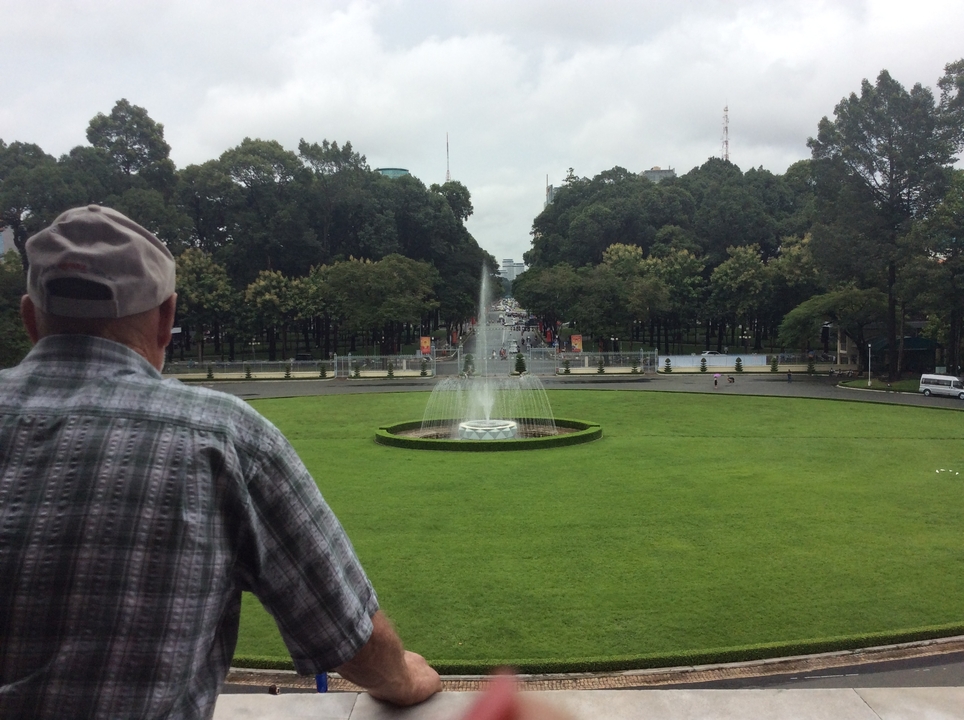 Homme âgé surplombant une fontaine dans un parc avec un large boulevard.