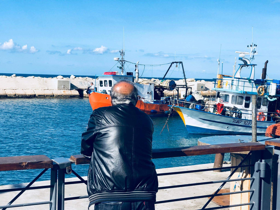 Man in leather jacket leans on a railing looking over fishing boats.