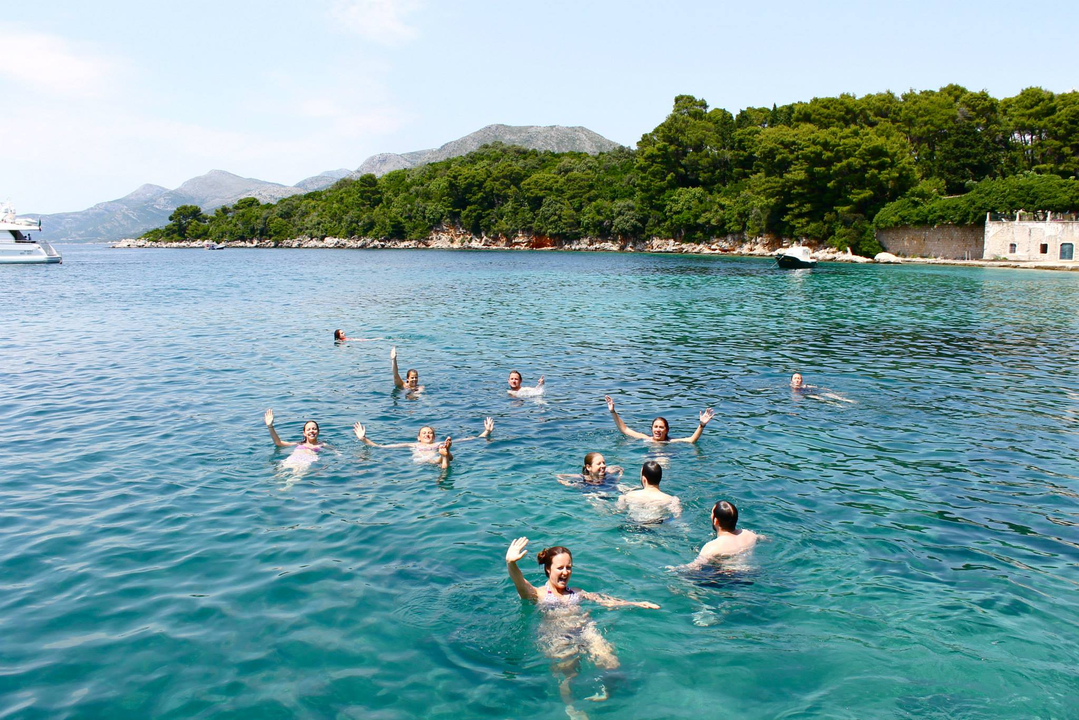 Personnes nageant dans une eau bleue claire avec un rivage boisé en arrière-plan.
