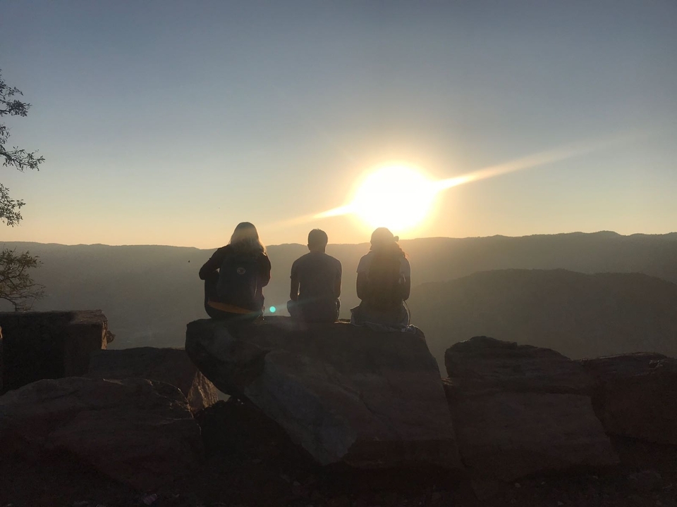 Trois personnes sur un rocher, silhouettes contre le soleil couchant.