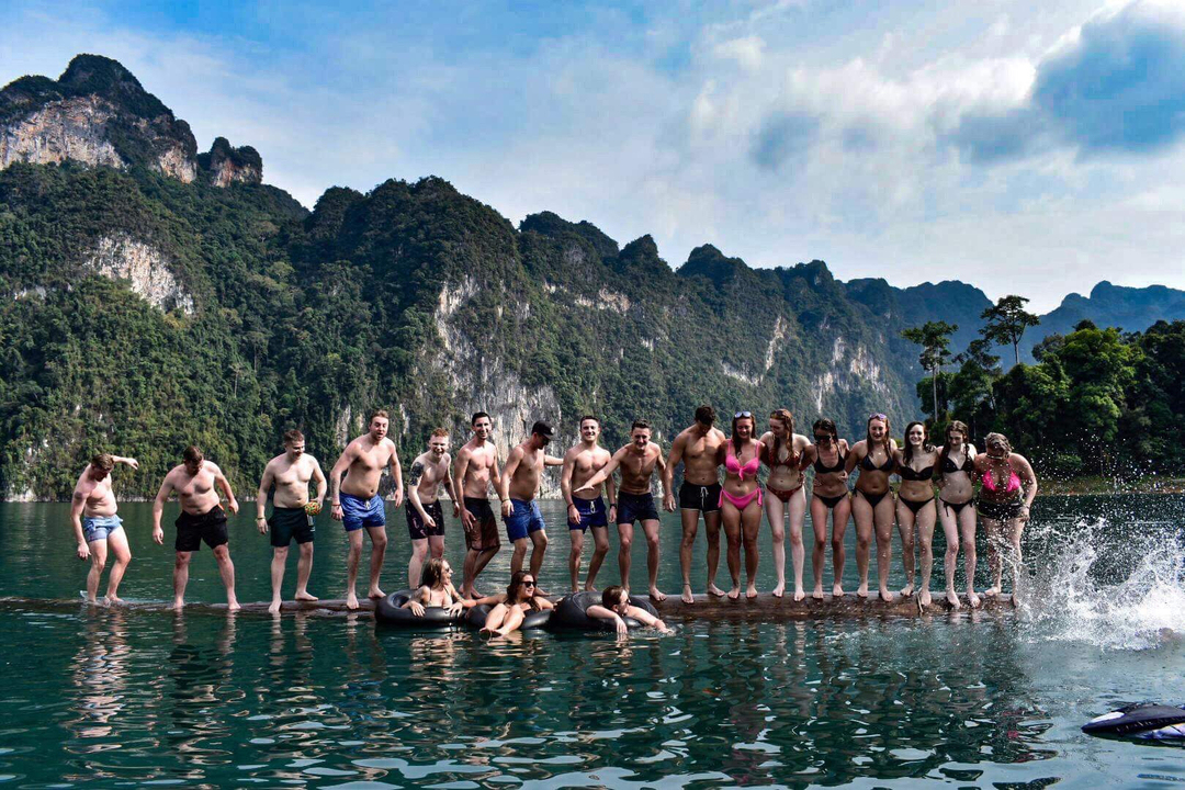 Group of people standing on log in water with mountainous backdrop.
