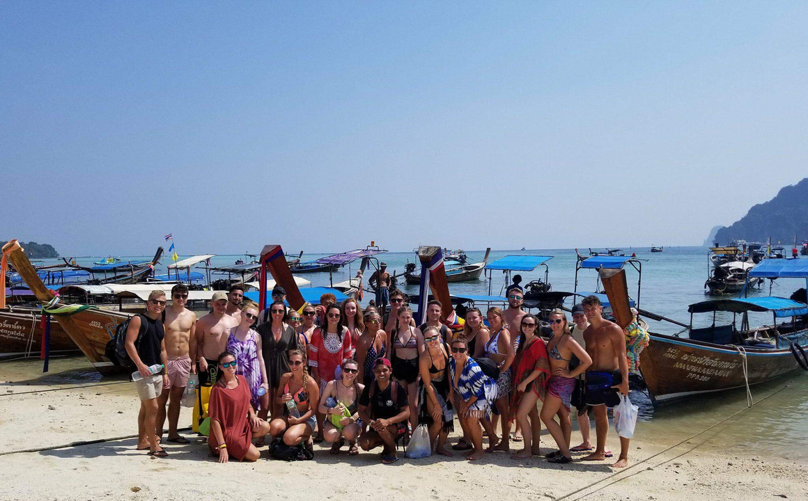 Group photo on a sandy beach with longtail boats in the background.