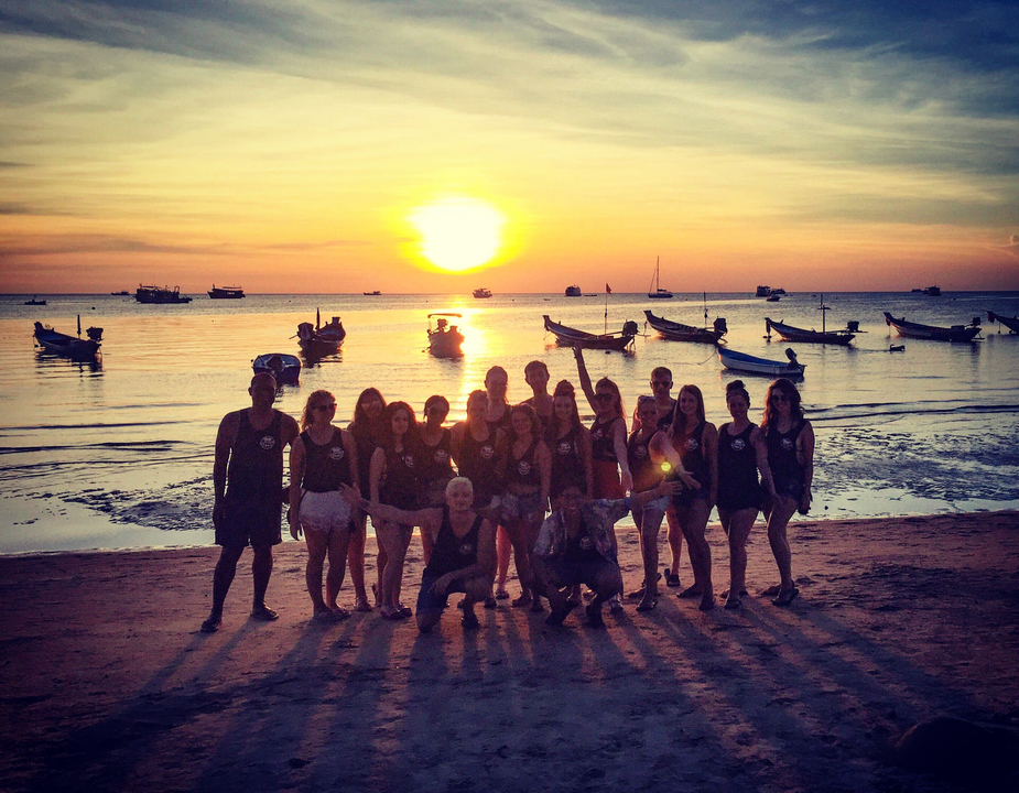 Group of people in front of a sunset at the beach with boats in the water.