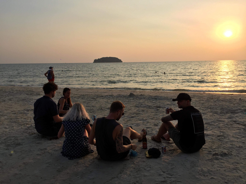 Group of people sitting on a beach at sunset with an island in the distance.