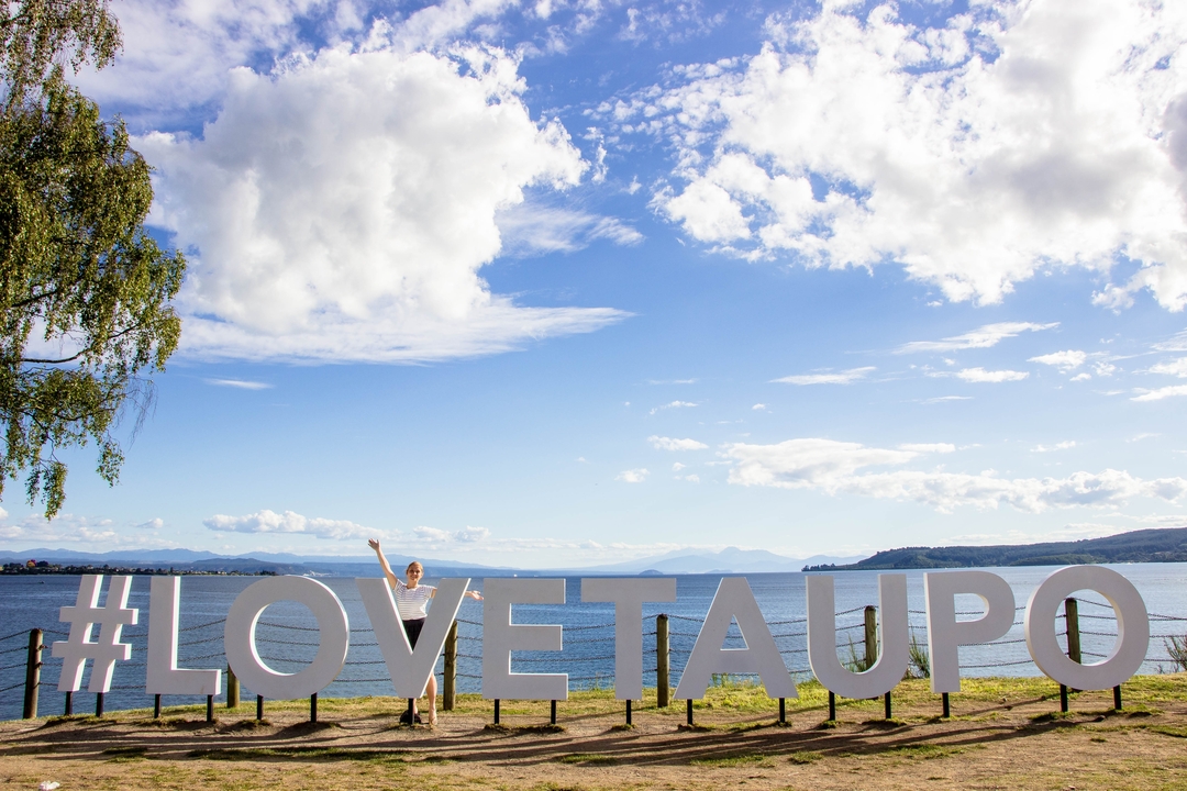 Personne posant avec le panneau #LOVETAUPO au bord d'un lac.