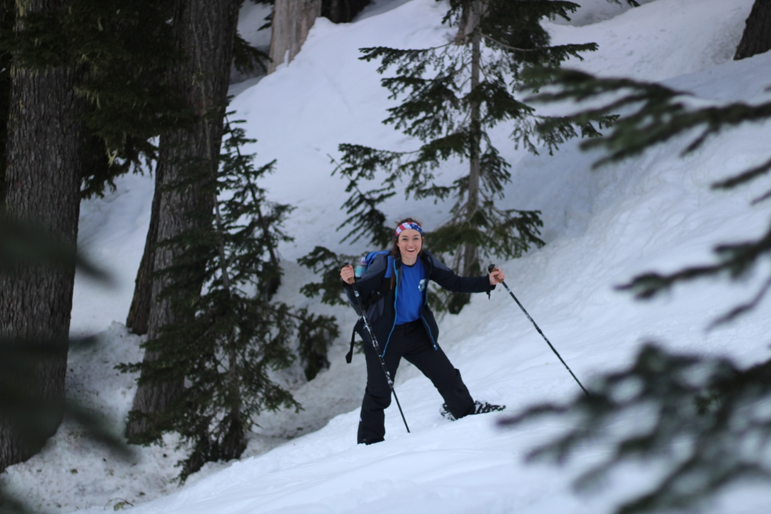 Une personne souriante avec un équipement de ski dans une forêt enneigée.