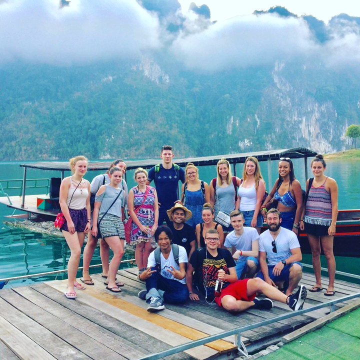 Large group of people posing by a lake with a boat in the background.