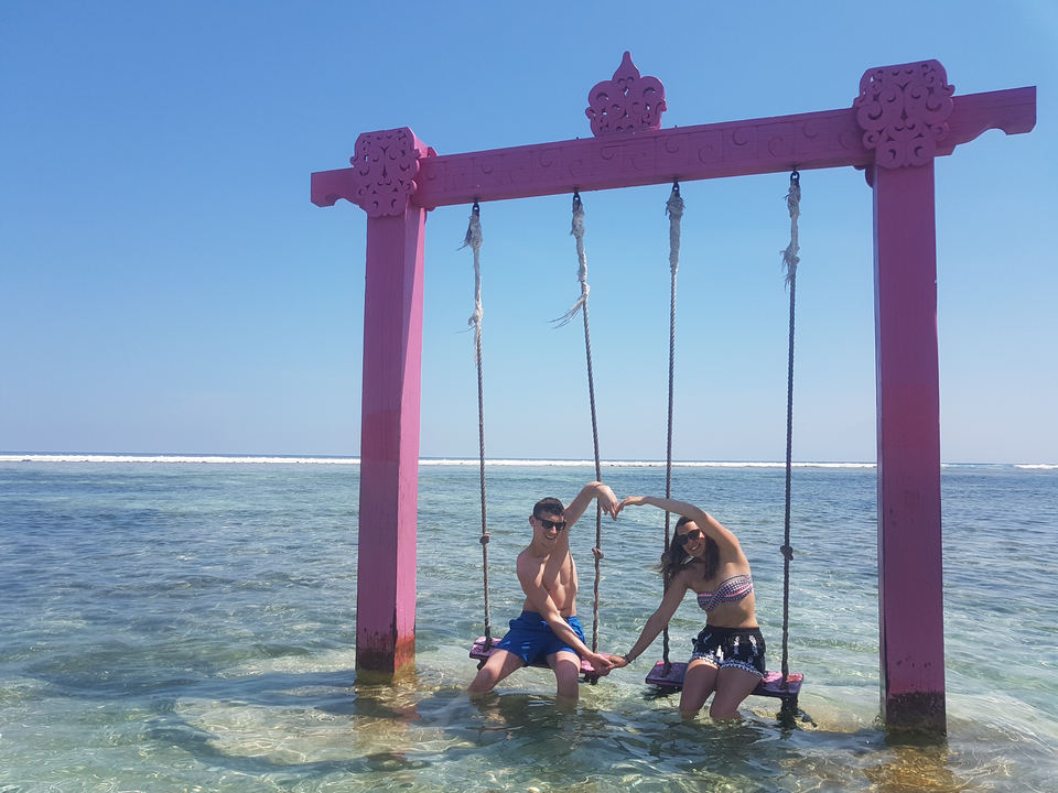 Two people sitting on a swing in shallow water making a heart shape with their hands.