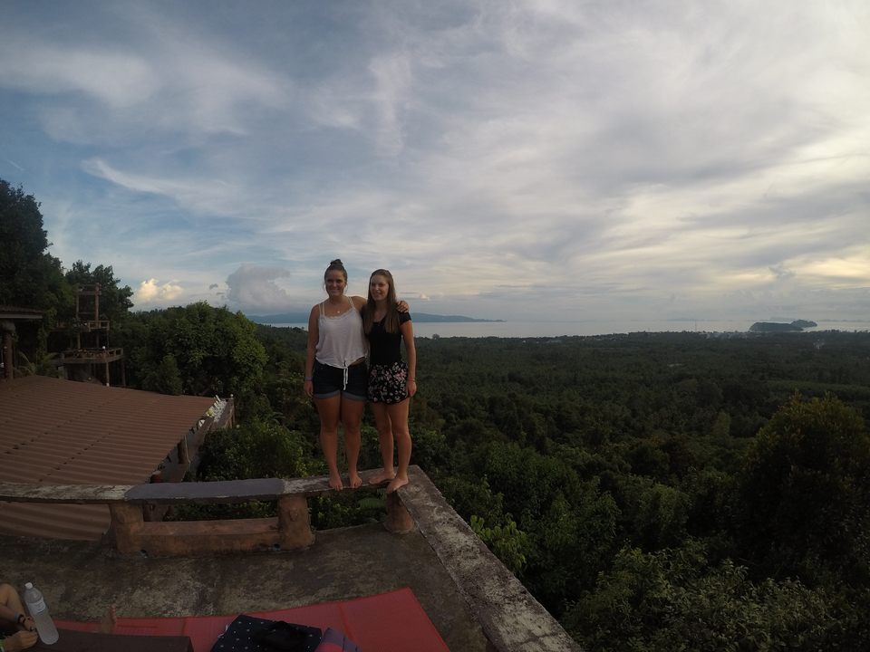 Two people standing on a ledge with a view of forested landscape and ocean.
