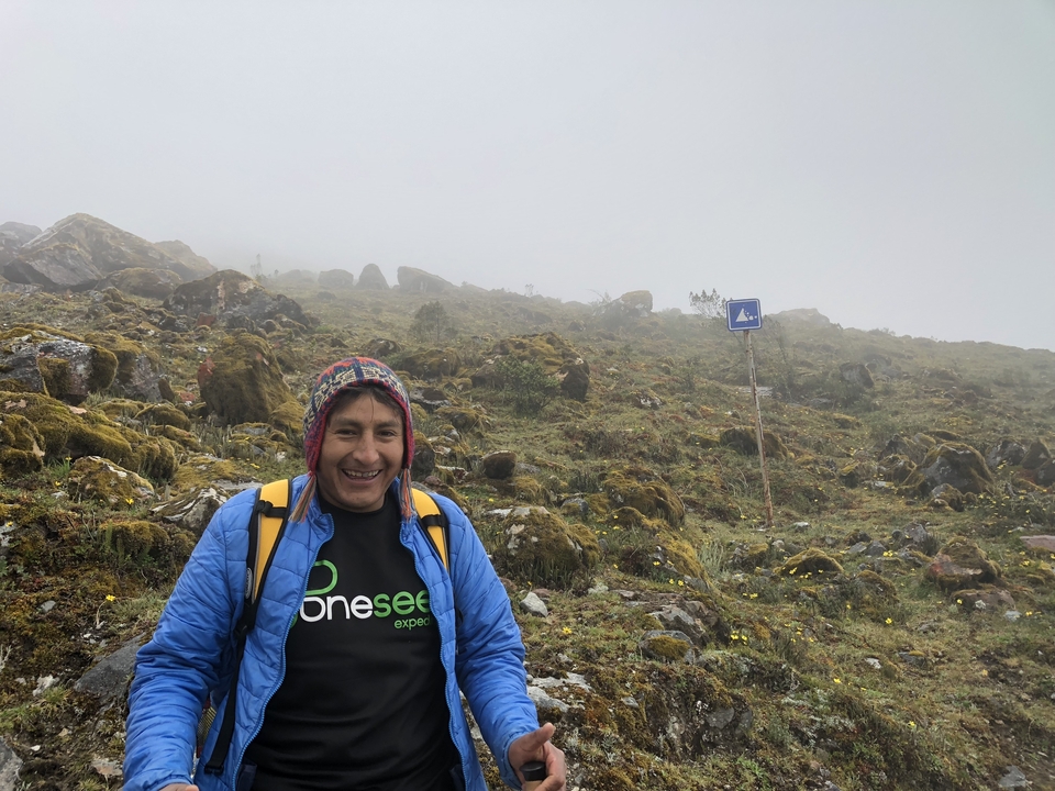 Homme en tenue de trekking souriant avec une colline brumeuse derrière lui.