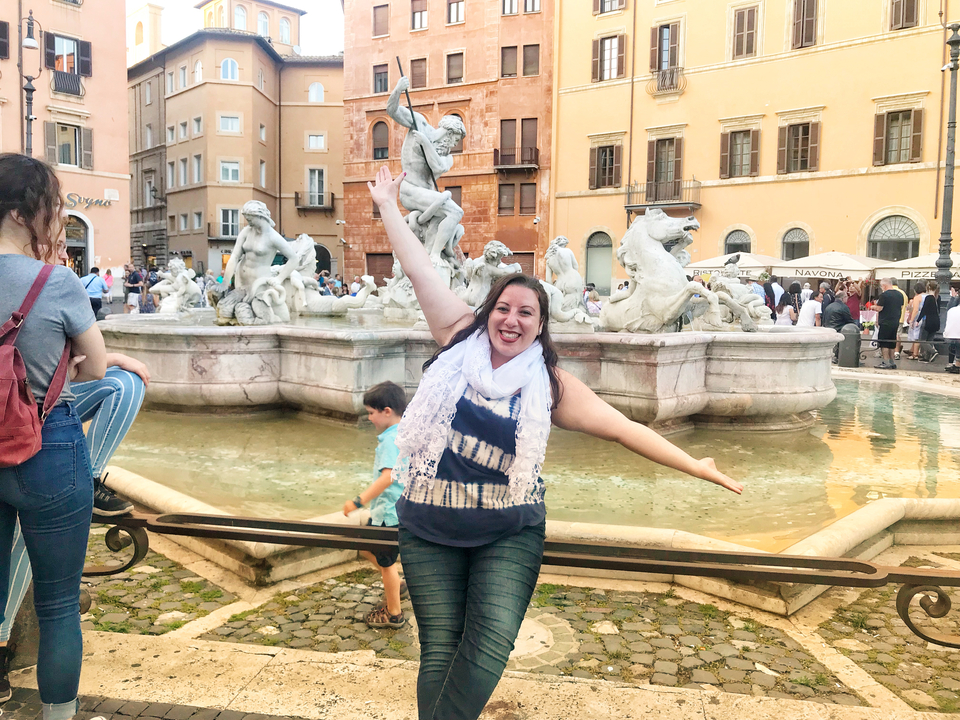 Femme souriante assise au bord d'une fontaine avec des statues sur une place publique.