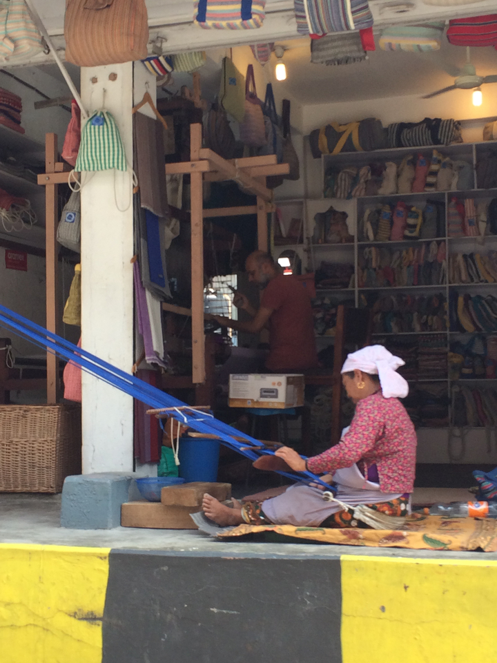 Scène d'intérieur d'un atelier d'artisanat avec deux personnes, l'une tissant et l'autre à un bureau.