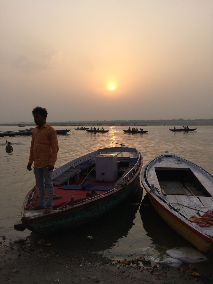 Personne debout près de bateaux sur une rivière au coucher du soleil, avec d'autres personnes en arrière-plan.
