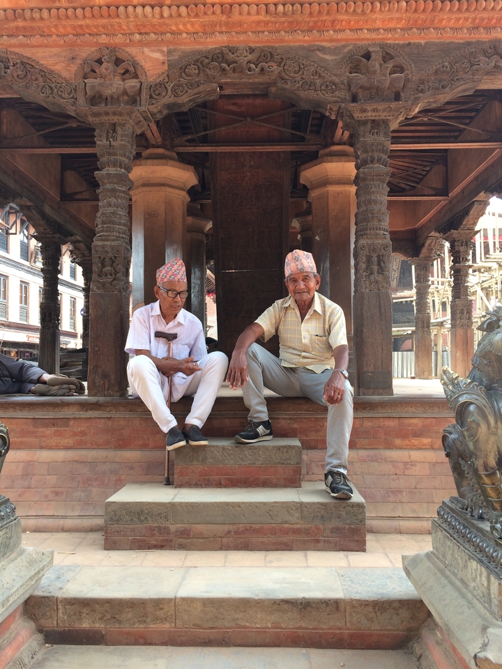 Deux hommes âgés assis dans un cadre traditionnel, coiffés d'un chapeau et adossés à un poteau en bois sculpté.