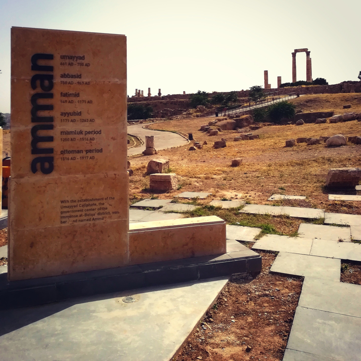 Stone inscription detailing the history of Amman with ruins in the background.