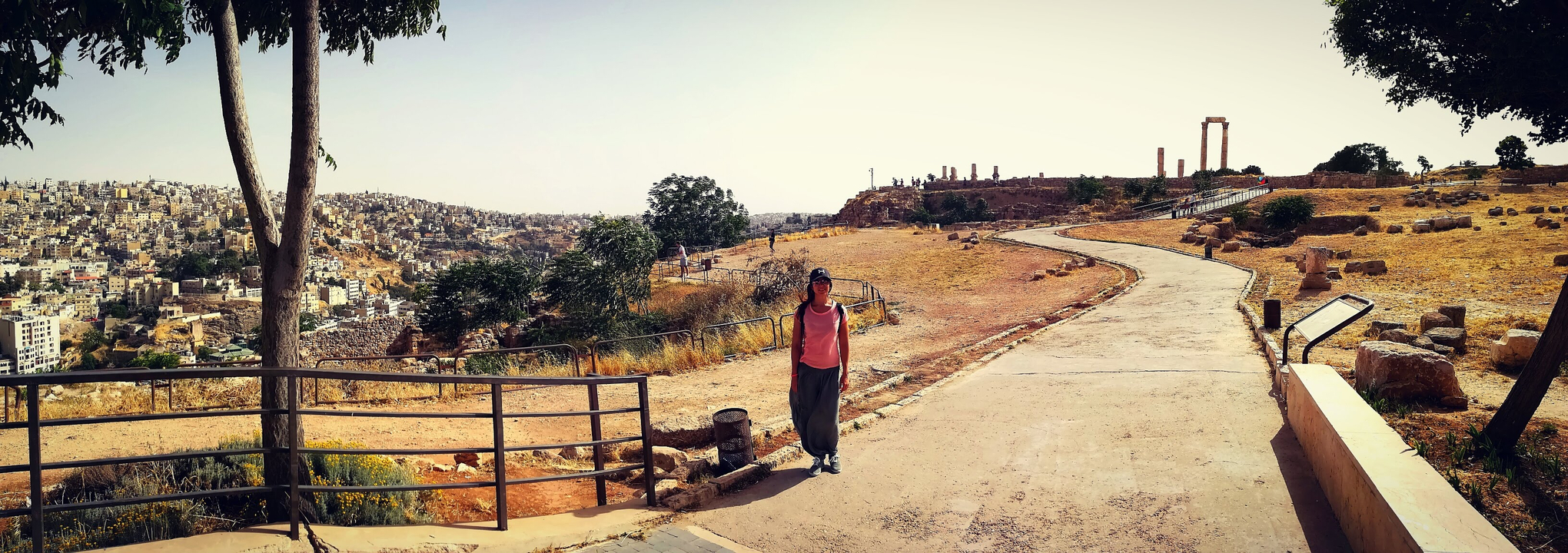 Femme marchant sur un chemin avec des ruines anciennes et une ville au loin.