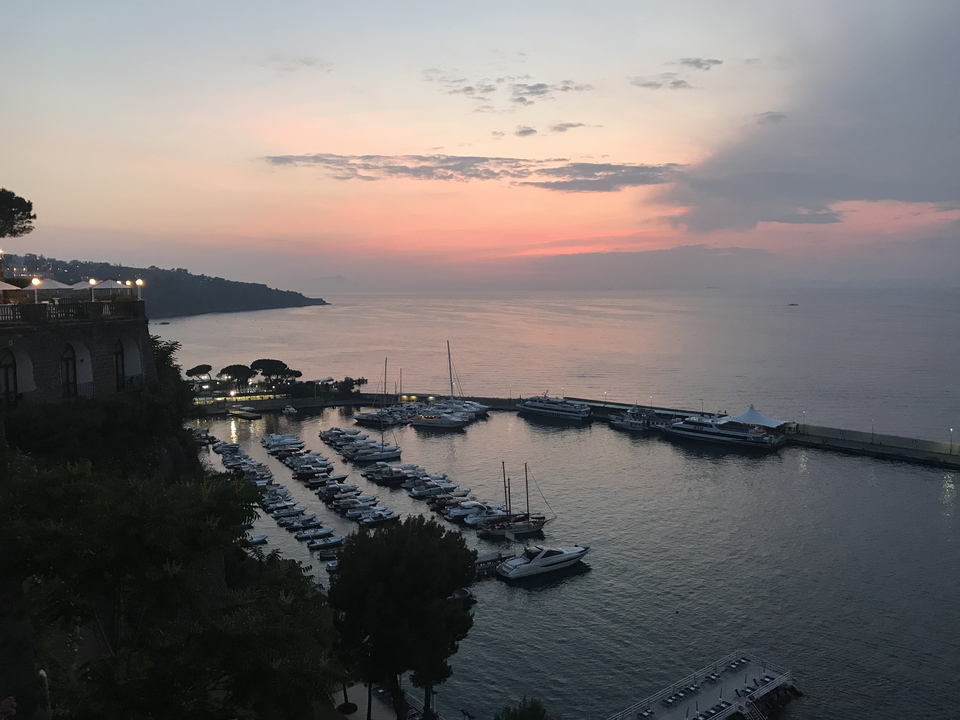 Coucher de soleil sur un port de plaisance avec des yachts et des collines au loin.