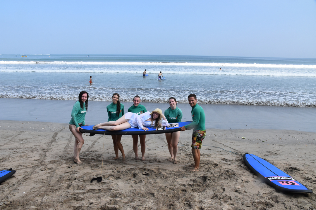 Group of six people posing on a surfboard on the beach.