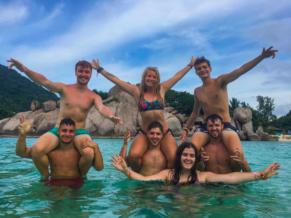 Group of people standing in the ocean, some on shoulders, under blue skies.
