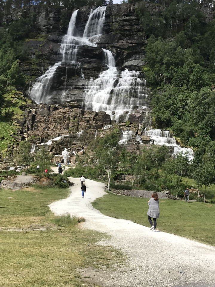Personnes marchant devant une cascade dans une région luxuriante.
