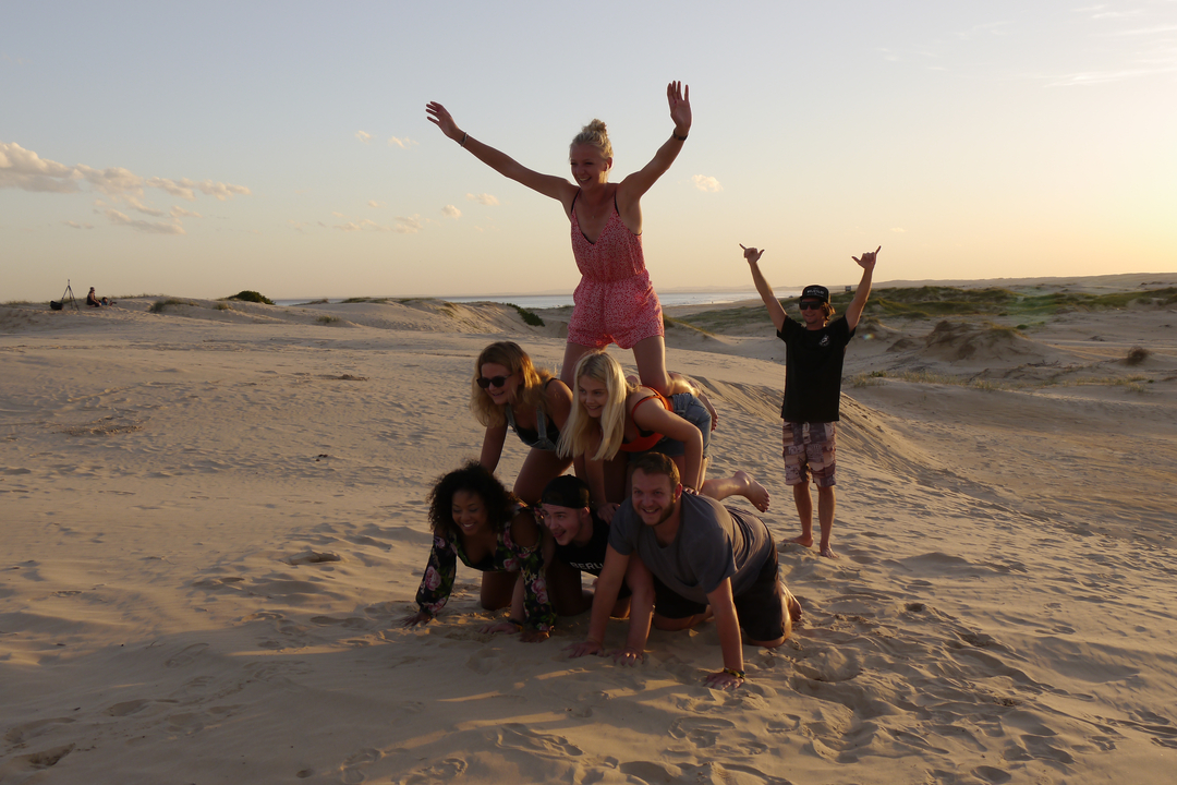 Group of people forming a human pyramid on sandy dunes during sunset.