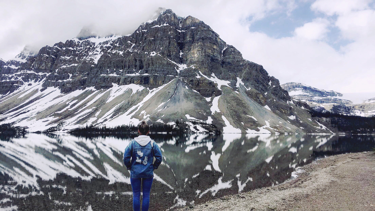 Eine Person steht ehrfürchtig vor einem Berg, der sich in einem See spiegelt.