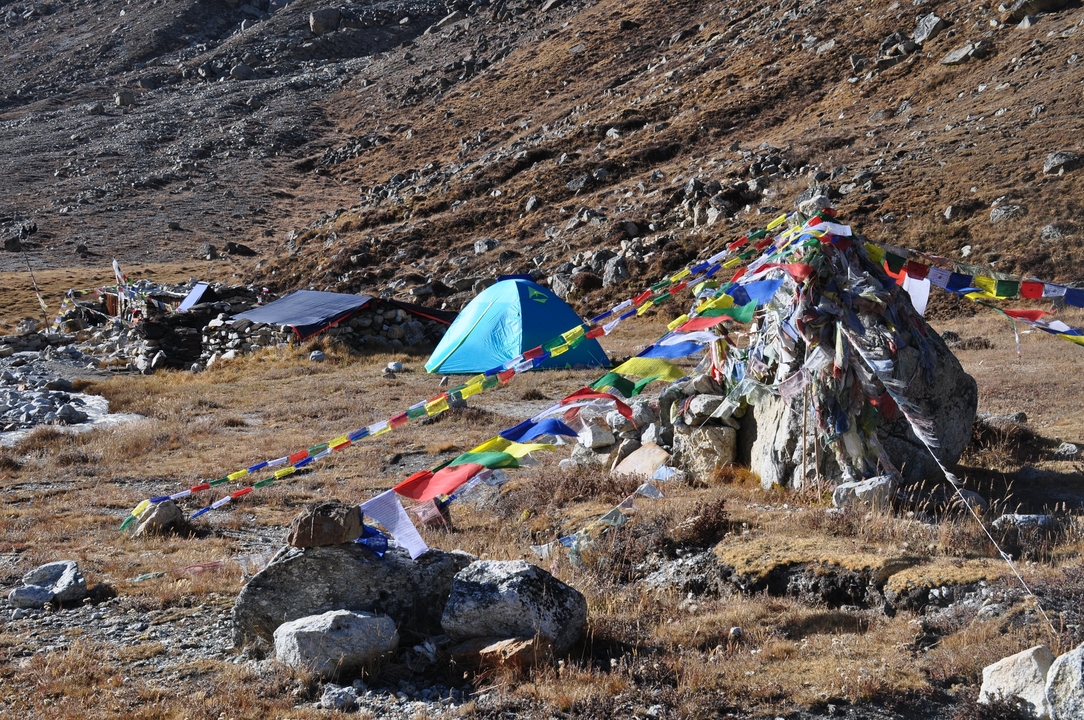 A lone blue tent sits beside fluttering prayer flags on a rugged alpine meadow.