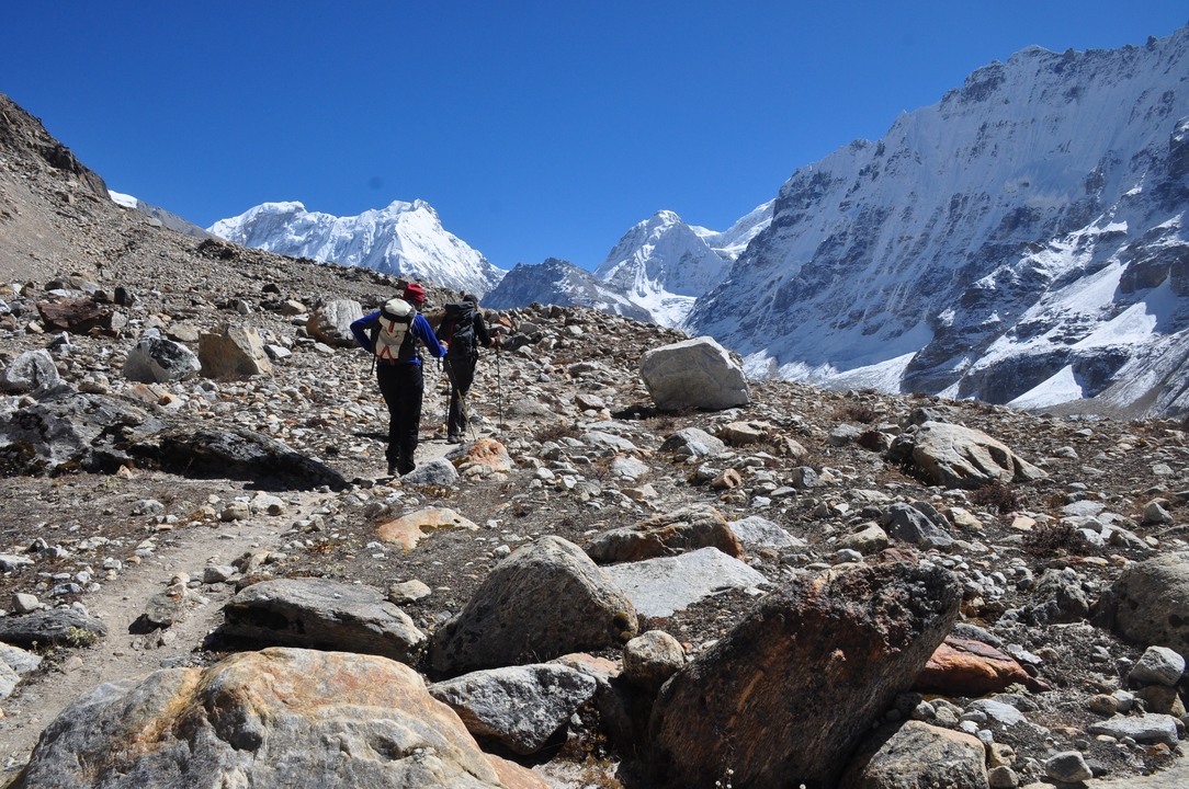 Trekkers with backpacks make their way along a rocky trail beneath towering ice-capped peaks.