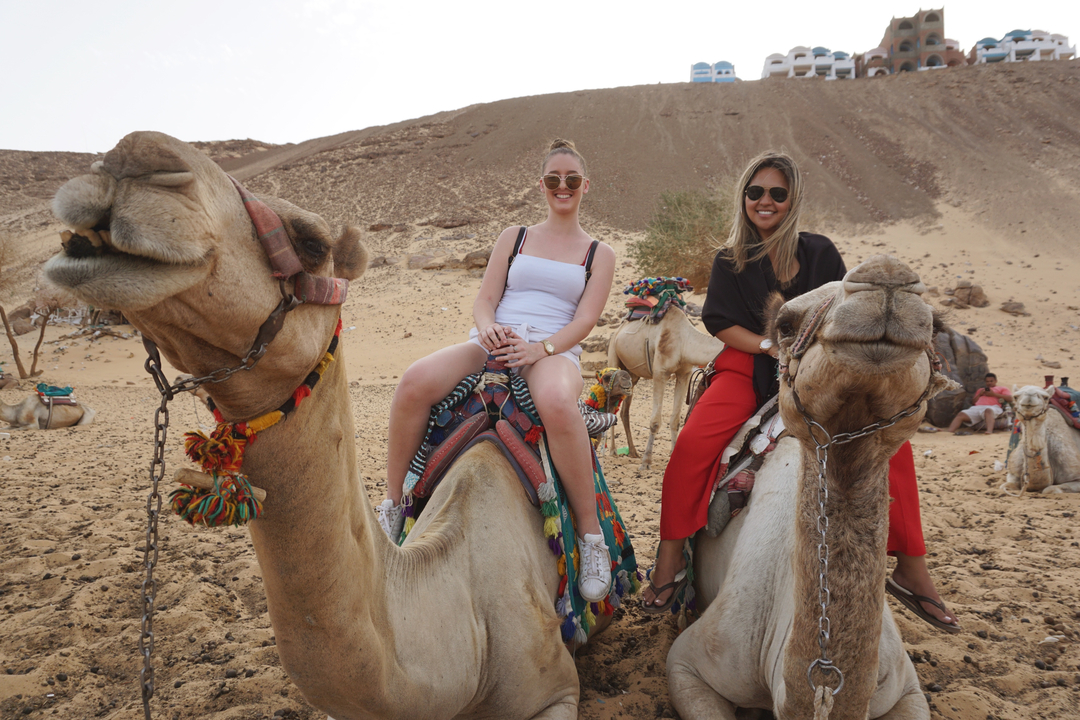 Two women riding camels in a desert landscape with hills in the background.