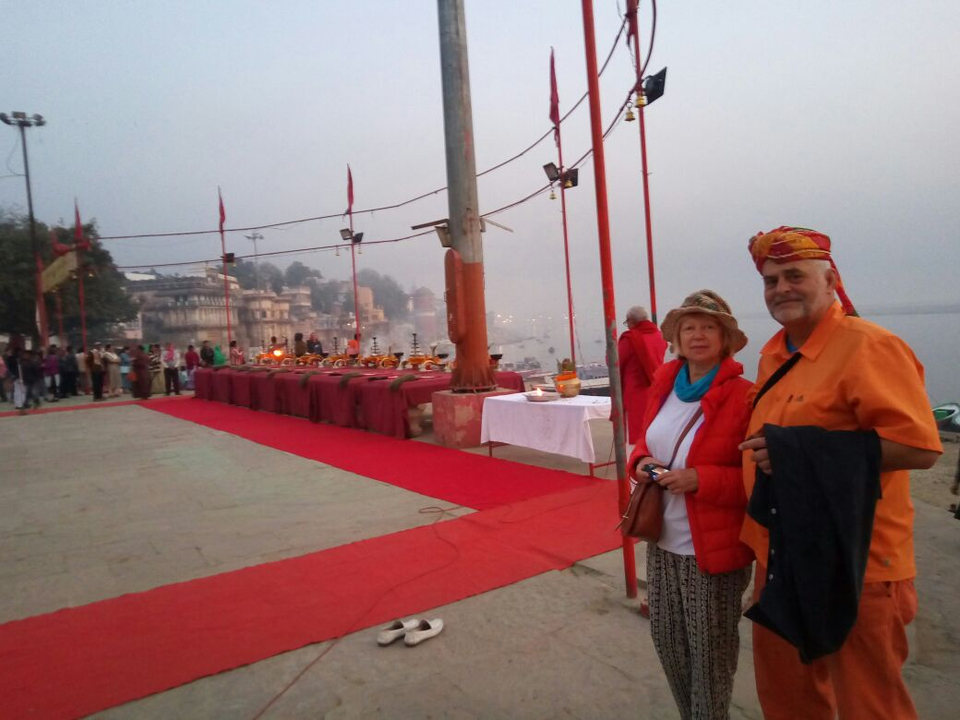 Travellers stand near a red-carpeted setup for the evening Ganga Aarti ritual on a misty riverside.