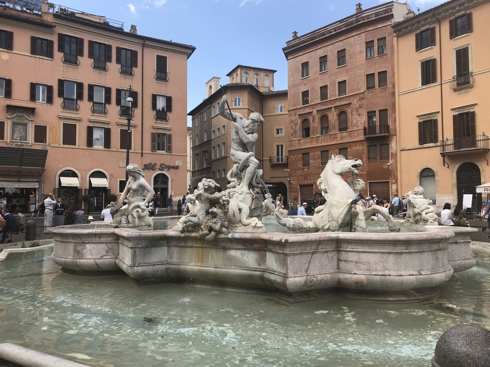 Fontaine sur une place romaine entourée de bâtiments.