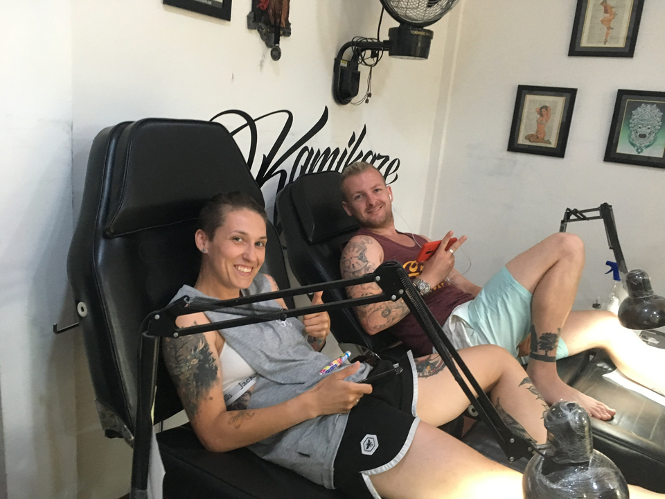 Two people with tattoos sitting on chairs, giving thumbs up.