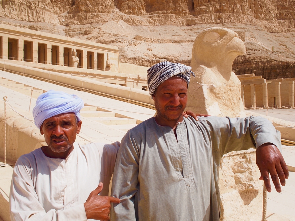Deux hommes en tenue traditionnelle posant avec des statues anciennes.
