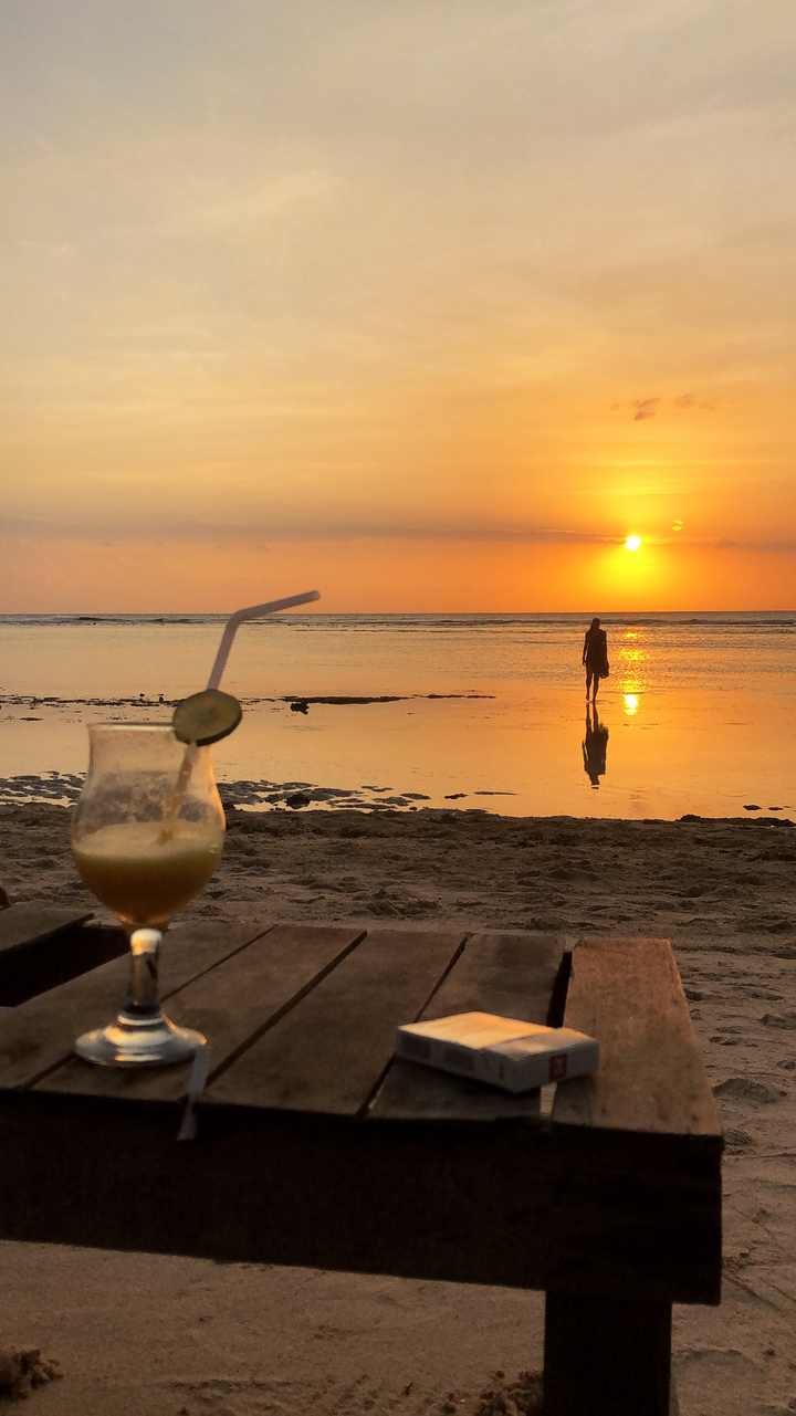 Sunset on a beach with a cocktail in the foreground and a person walking.