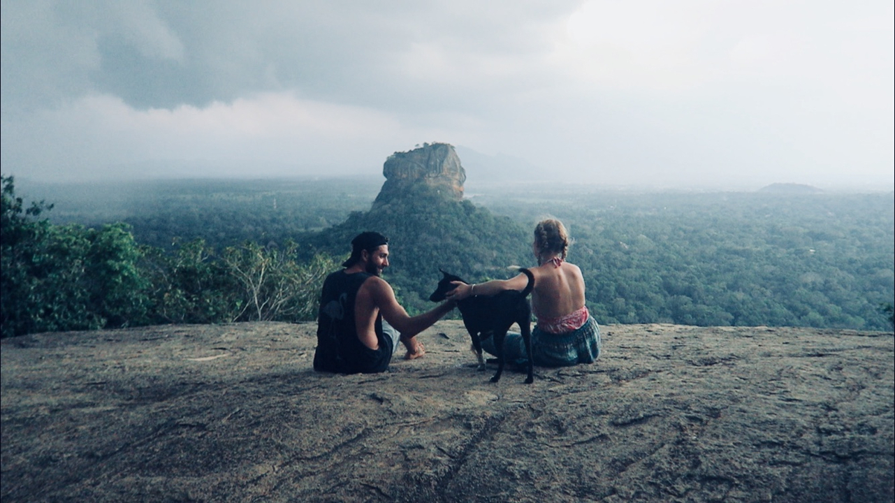 Deux personnes avec un chien assises sur une colline rocheuse avec une vue sur le rocher de Sigiriya.