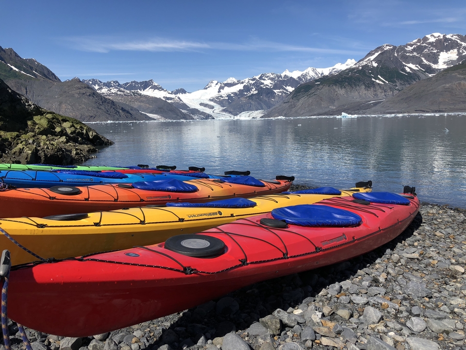 Kayaks colorés sur un rivage rocheux avec des montagnes enneigées en arrière-plan.