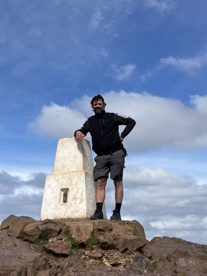 Hombre de pie sobre rocas junto a un vértice geodésico contra un cielo nublado.