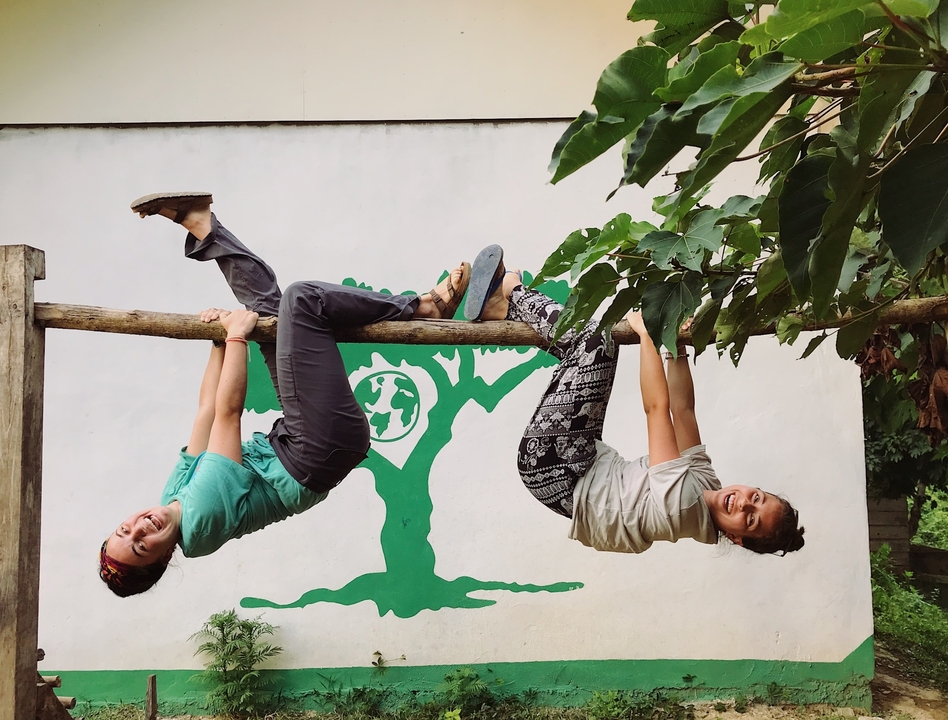 Two people playfully hanging from a wooden bar with a mural behind.